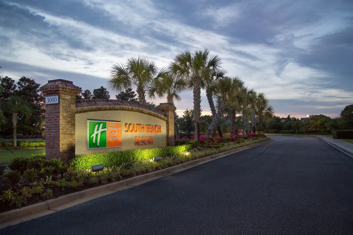 A warmly illuminated resort sign stands at the landscaped entrance, surrounded by palm trees and well-kept gardens. The lighting suggests an inviting atmosphere for guests arriving in the evening.