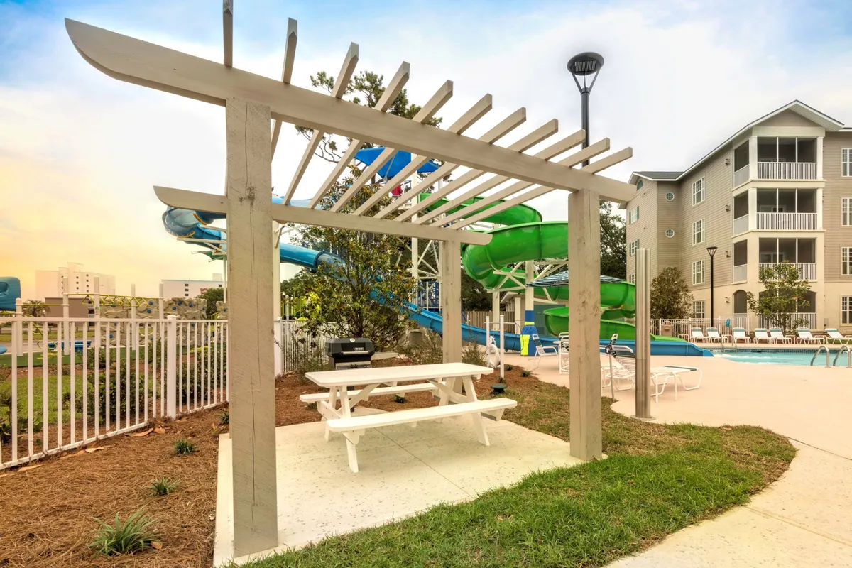 An inviting pool scene that combines water slides, a picnic table, lush greenery, and a sparkling swimming pool. Families gather for picnics and games, while others relax poolside or enjoy the water. The setting is relaxed yet playful, underscoring opportunities for socializing and creating summer memories.