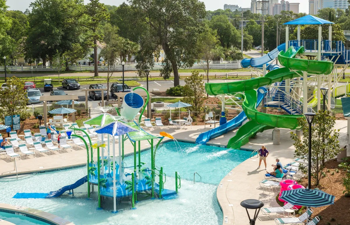 A vibrant outdoor pool area featuring colorful water slides, a splash pad, a spacious swimming pool, and plenty of lounge chairs. Children are seen playing and laughing, while families gather by the edge of the water. The pool is active, exuding a joyful and energetic vibe, with the central focus on family water fun.