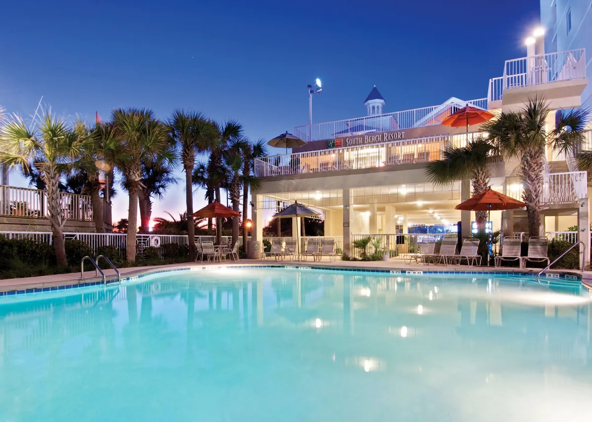 An inviting outdoor scene featuring the resort's swimming pool surrounded by lush palm trees, sun umbrellas, and the elegant building facade. The relaxed atmosphere is accentuated by guests lounging poolside, with hints of sunset colors reflecting on the water, suggesting a romantic and social ambiance as evening falls.