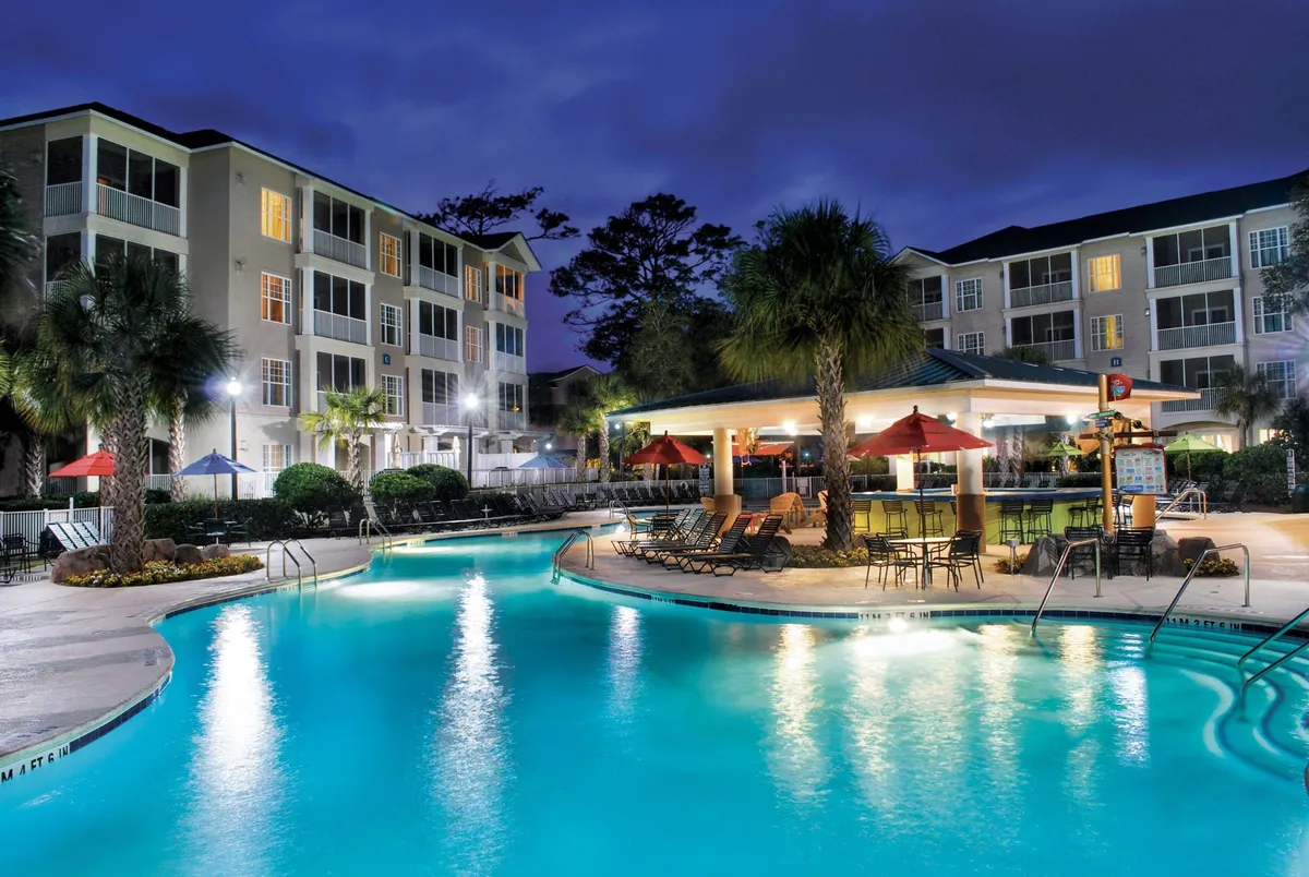 An elegant outdoor pool scene featuring a stylish poolside bar (Captain Cliff’s), lounge chairs, illuminated buildings, and palm trees. Guests relax by the water and enjoy cocktails at sunset, evoking a festive yet laid-back adult retreat.